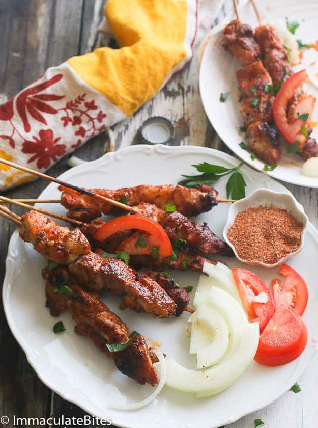chicken suya skewers on a white plate with onions and tomatoes.