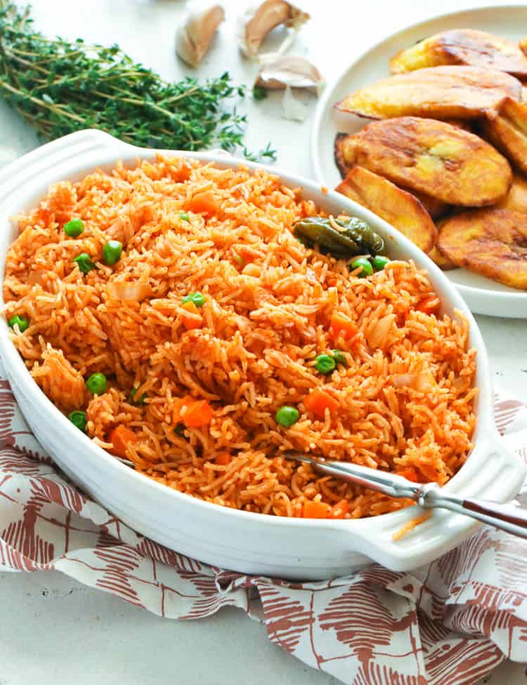 Jollof Rice in a casserole dish with fried sweet plantains and sprigs of thyme in the background.