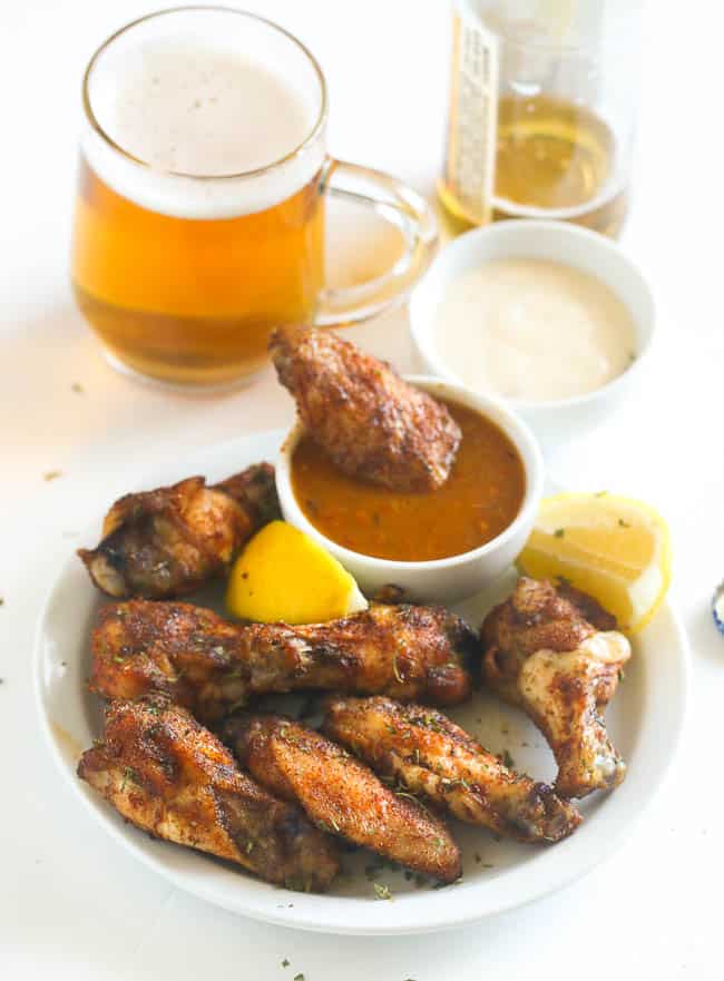 A Plate of Baked Jerk Chicken Wings with One Wing Dipped in a Sauce and Two Mugs of Beer in the Background