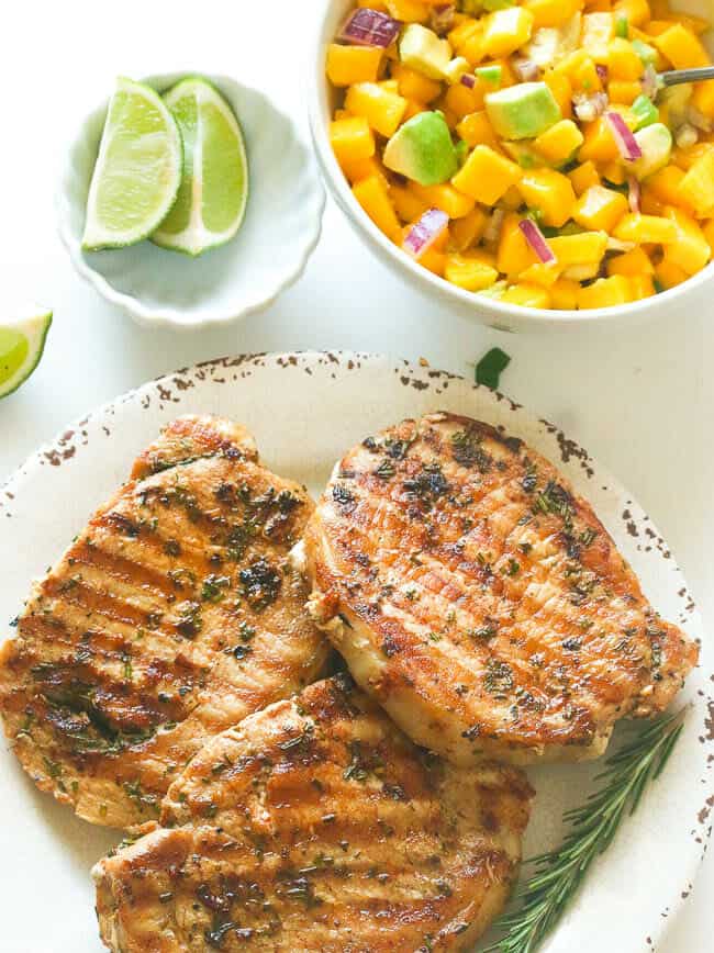 A Plate with Three Marinated Pork Chop Served with Mango Salsa and Lime Slices on the Side