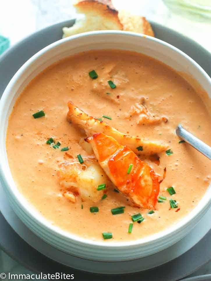 a white bowl of lobster bisque topped with lobster meat with crusty bread in the background