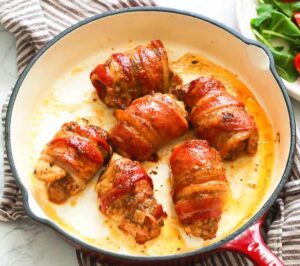 Bacon-wrapped chicken in a white skillet with a side salad in the background