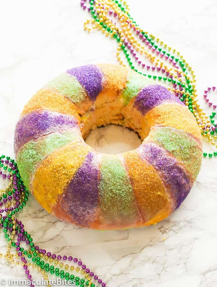 a whole king cake glazed with green, purple, and yellow with colorful beads in the background
