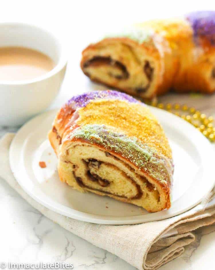 a slice of King Cake on a plate served with coffee in the background