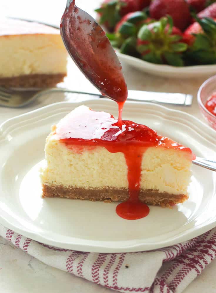 A slice of cheesy New York Cheesecake with strawberry sauce being drizzled over it