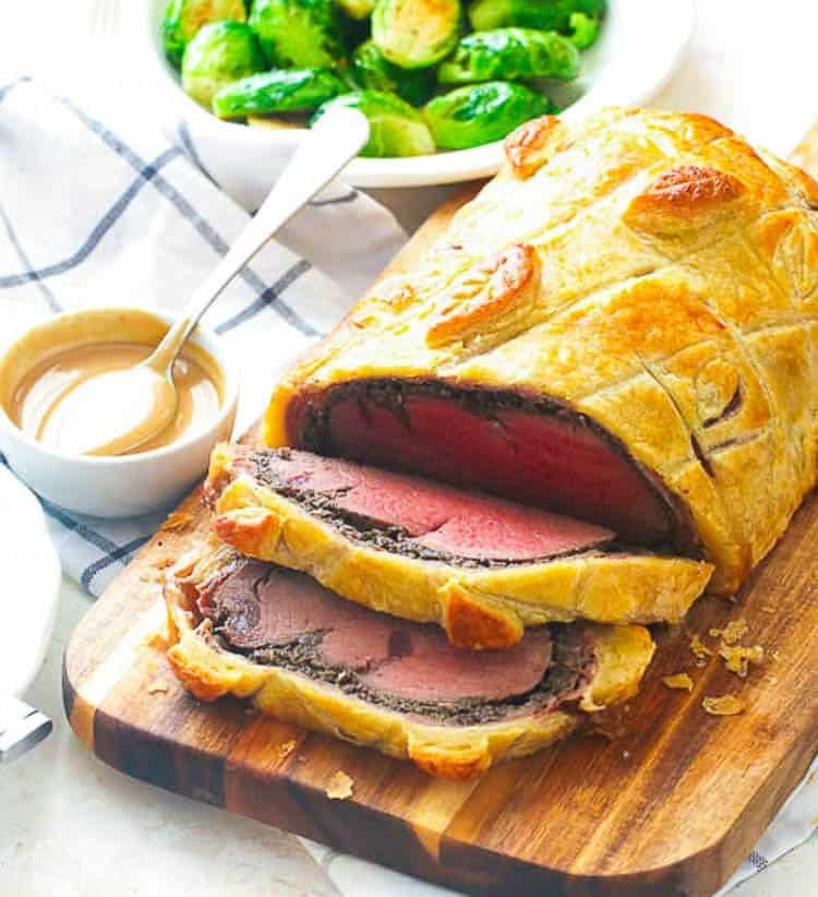 sliced Beef Wellington on a chopping board served with sauce and brussel sprouts on the side