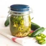 Pickled jalapenos in a jar with jalapeno peppers scattered around the jar