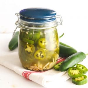 Pickled jalapenos in a jar with jalapeno peppers scattered around the jar