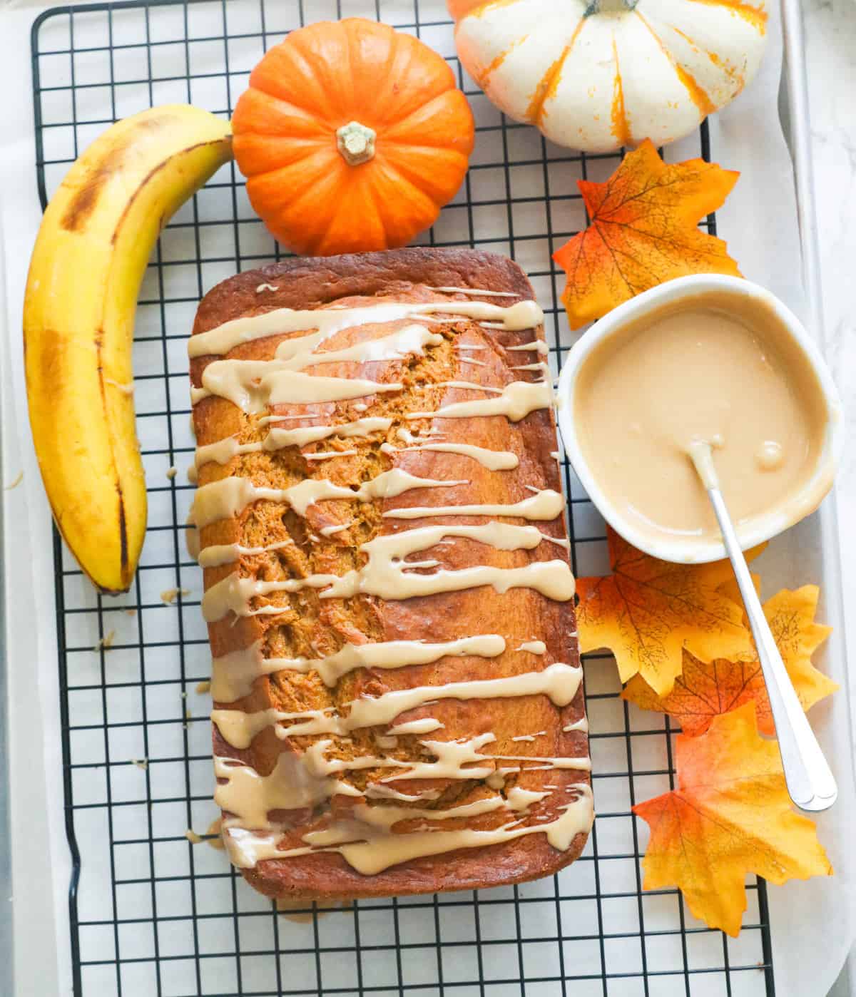 Pumpkin banana bread glazed in cooling rack with a whole banana and mini pumpkins beside it