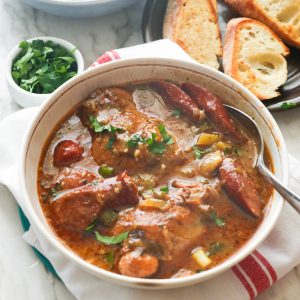 A bowl of chicken and sausage gumbo with parsley and garlic bread in the background