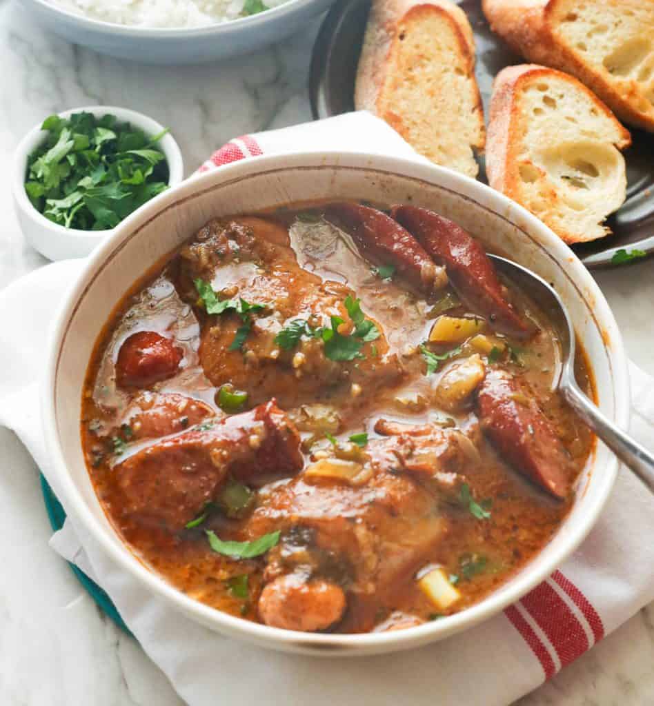 A bowl of chicken and sausage gumbo with parsley and garlic bread in the background