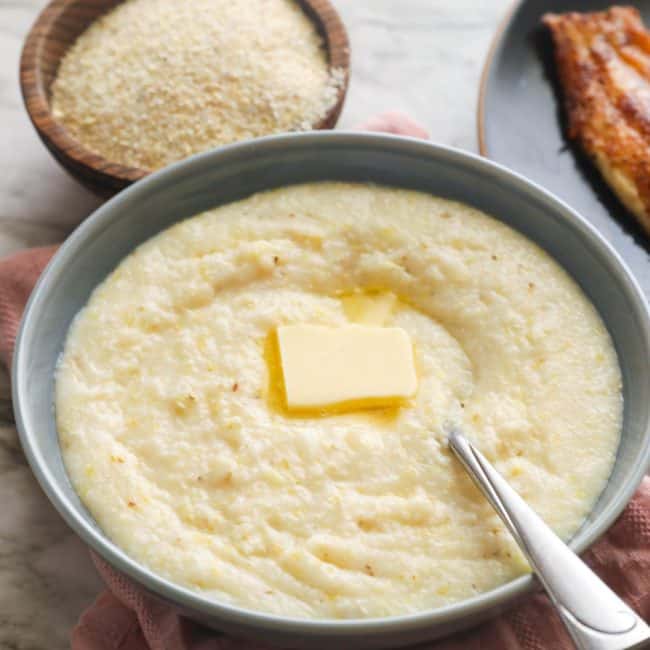 Stone-ground grits in a bowl topped with a pat of butter and blackened catfish in the background.