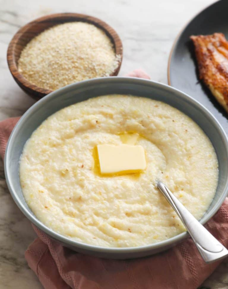 Stone-ground grits in a bowl topped with a pat of butter and blackened catfish in the background.