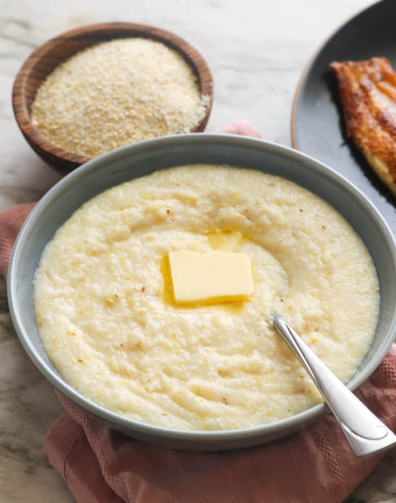 Stone-ground grits in a bowl topped with a pat of butter and blackened catfish in the background.