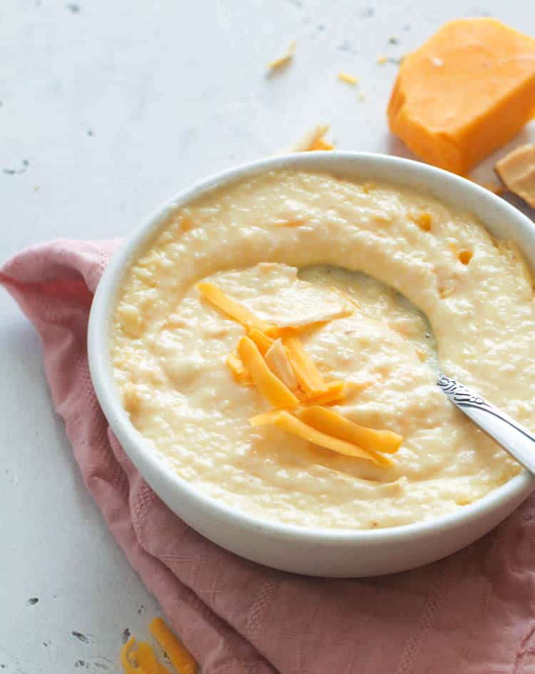 A bowl of creamy cheese grits topped with shaved cheese and a block of cheese in the background