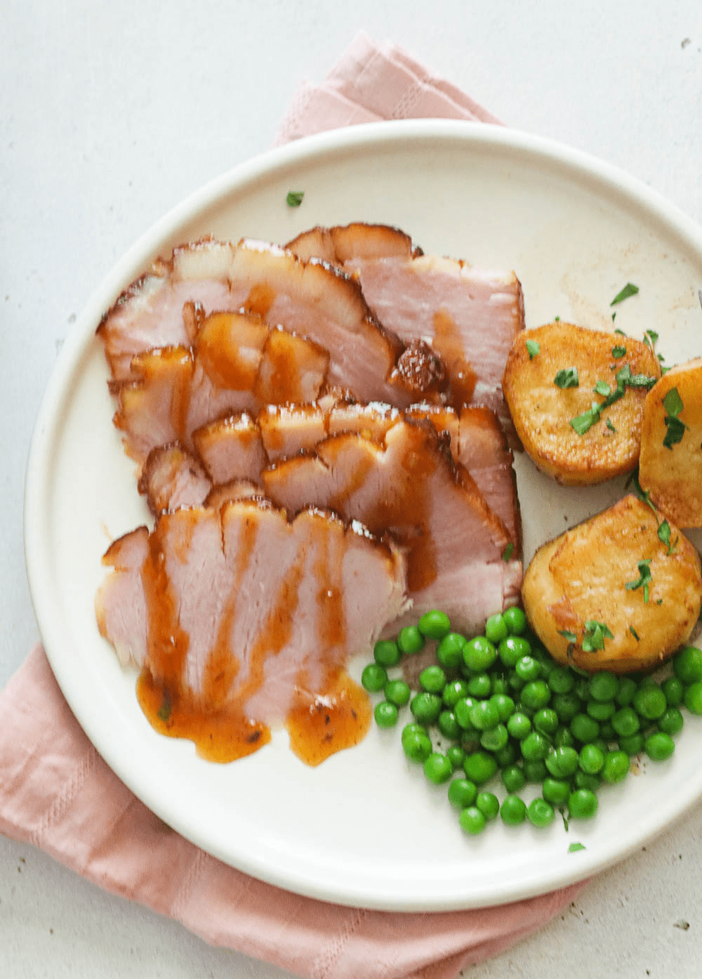 A plate of smoked ham slices drizzled with glaze and served with green peas and potatoes
