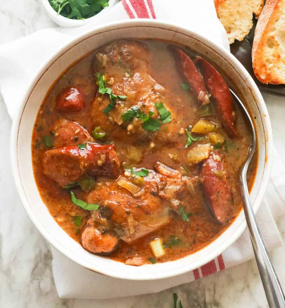 A bowl of chicken and sausage gumbo in a white bowl with homemade garlic bread in the background.