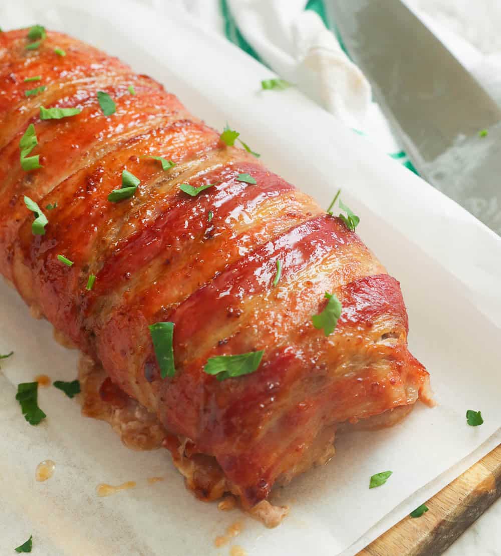 Bacon and meatloaf log on parchment paper over a wooden cutting board.