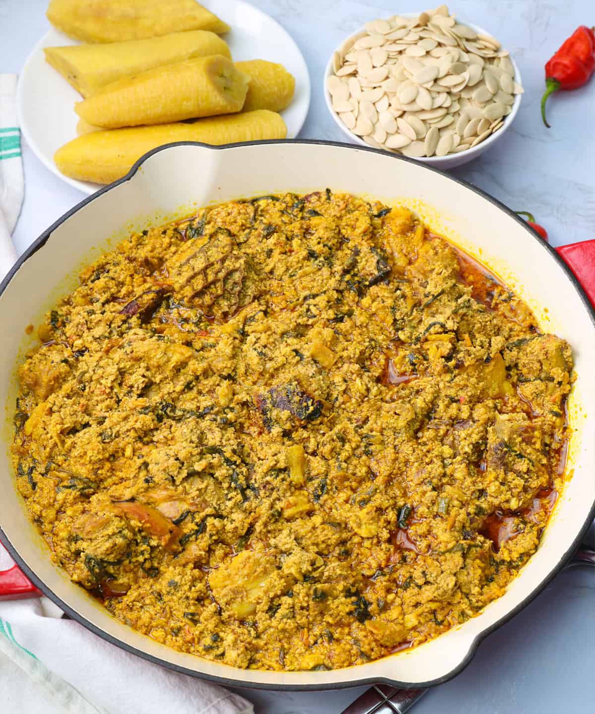 a pan of egusi soup with plantains and egusi seeds in the background