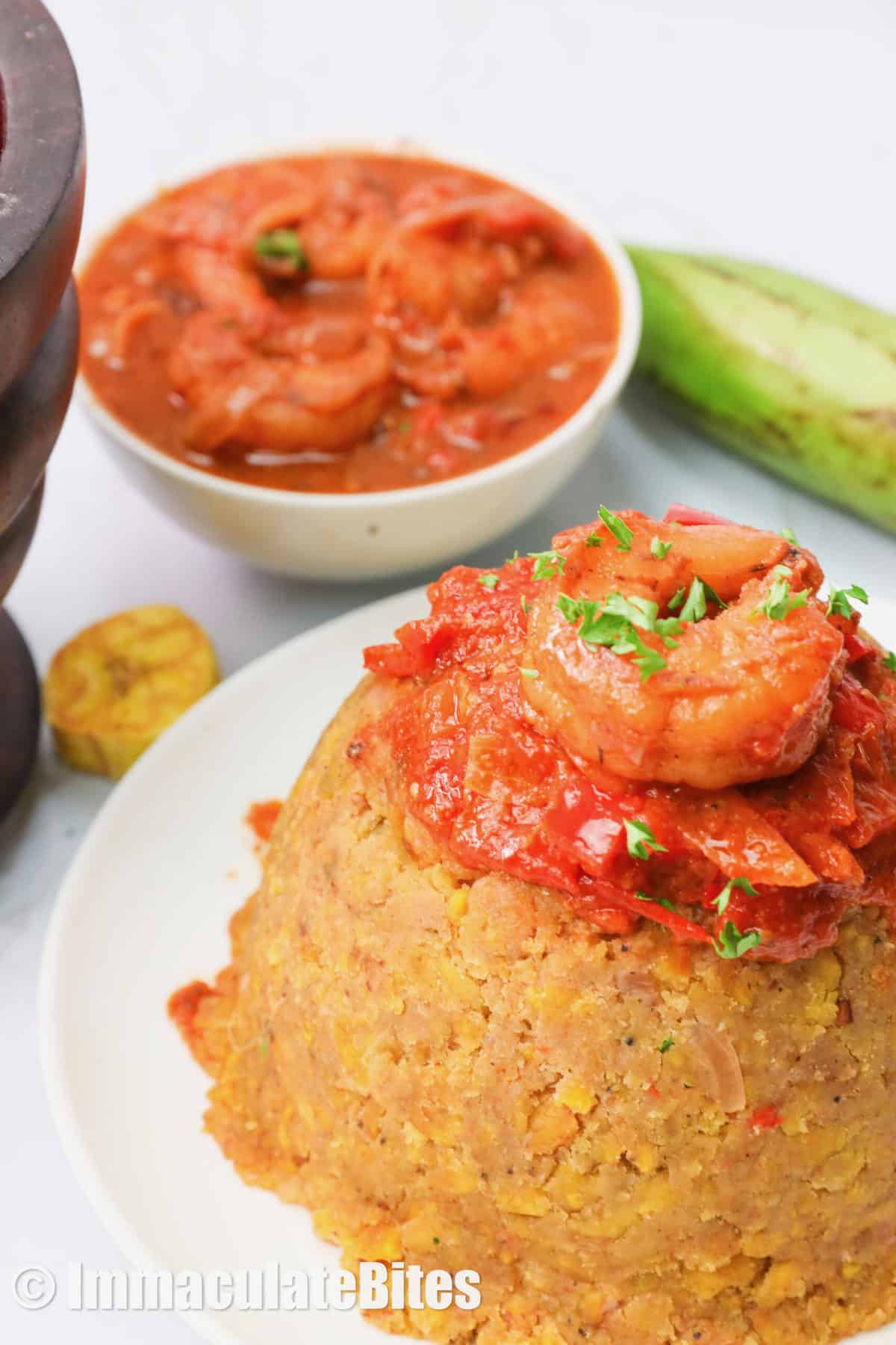Mofongo topped with shrimp sauce and more sauce and a plantain in the background