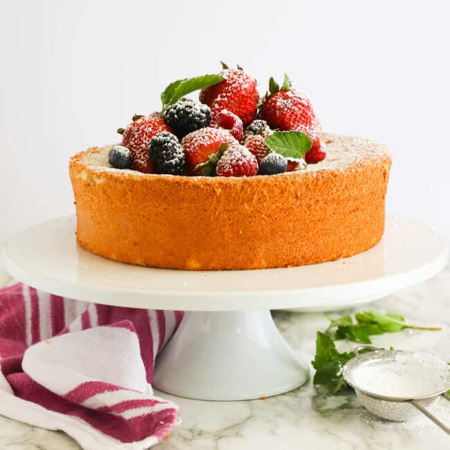 A whole angel food cake topped with fresh fruit on a white cake plate