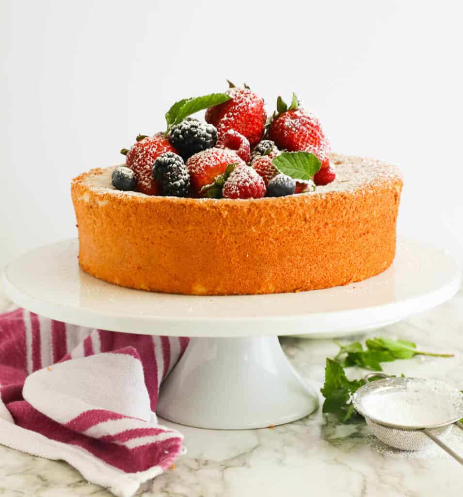 A whole angel food cake topped with fresh fruit on a white cake plate