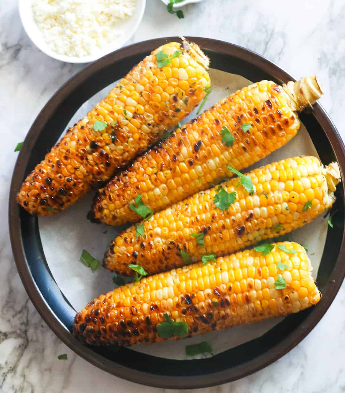 Grilled corn on a plate with cilantro on top and queso fresco on the side