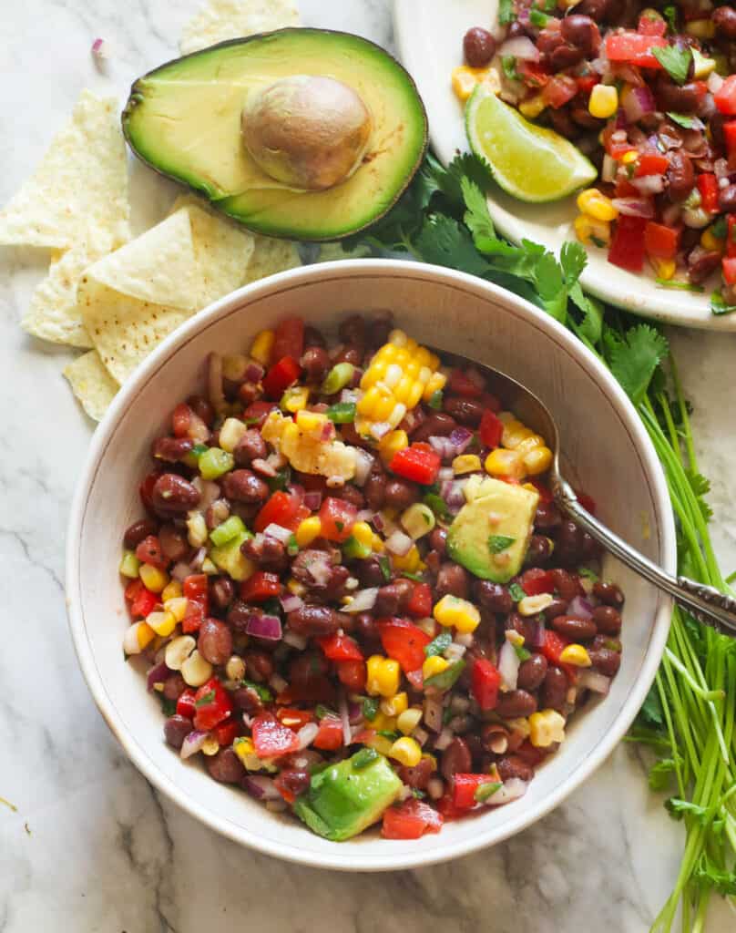 A bowl of black bean and corn salad with an avacodo and tortilla chips in the background