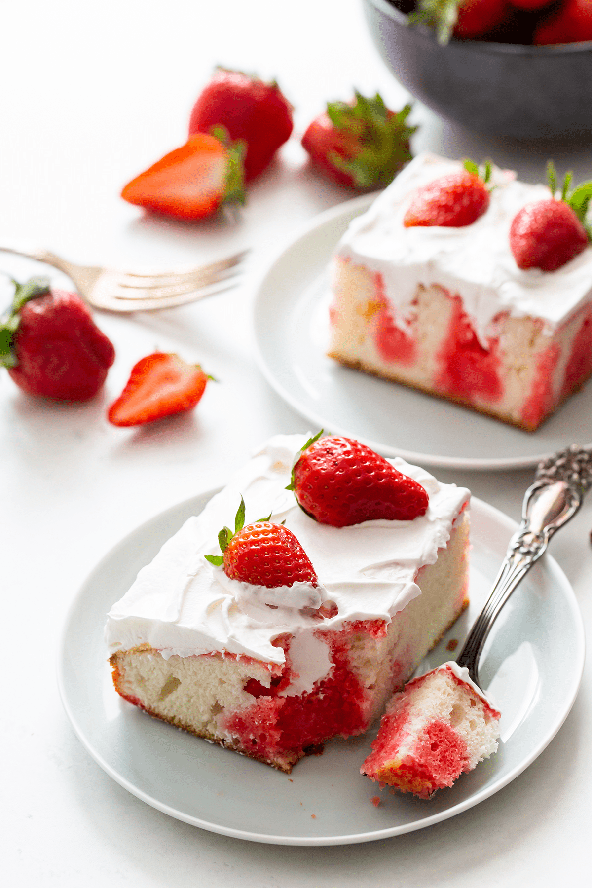 A square of strawberry poke cake on a white plate with fresh strawberries in the background