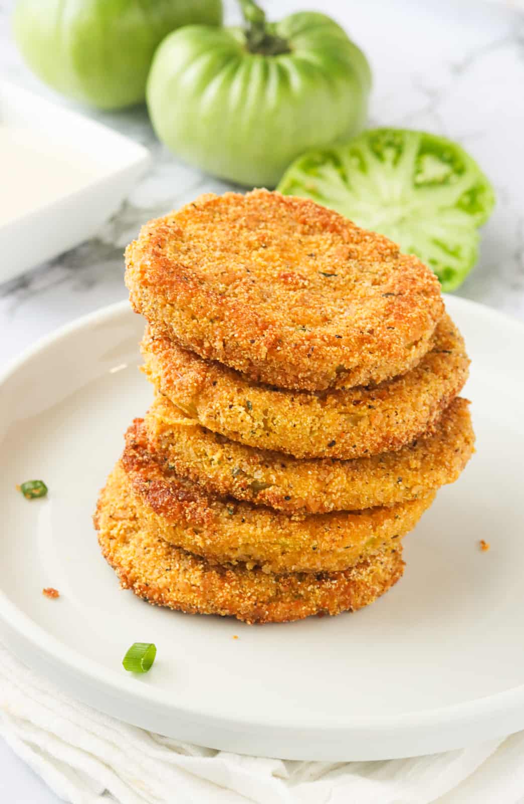 A stack of fried green tomatoes on a white plate with fresh green tomatoes in the background