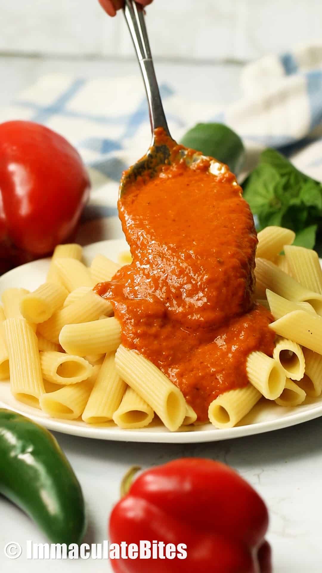 Drizzling Roasted Red Pepper Sauce over pasta with red peppers and jalapenos in the background