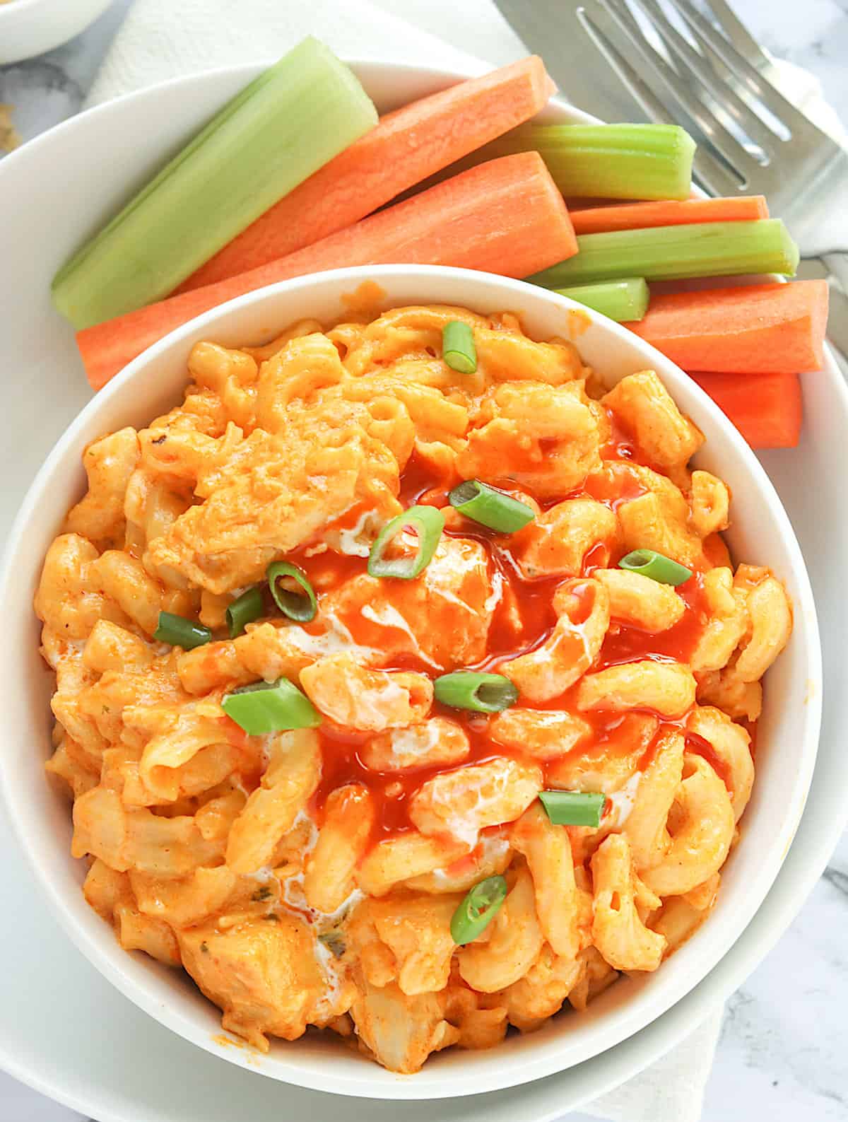 A tempting bowl of Buffalo Chicken Mac and Cheese drizzled with blue cheese dressing and hot sauce with celery and carrot sticks on the side