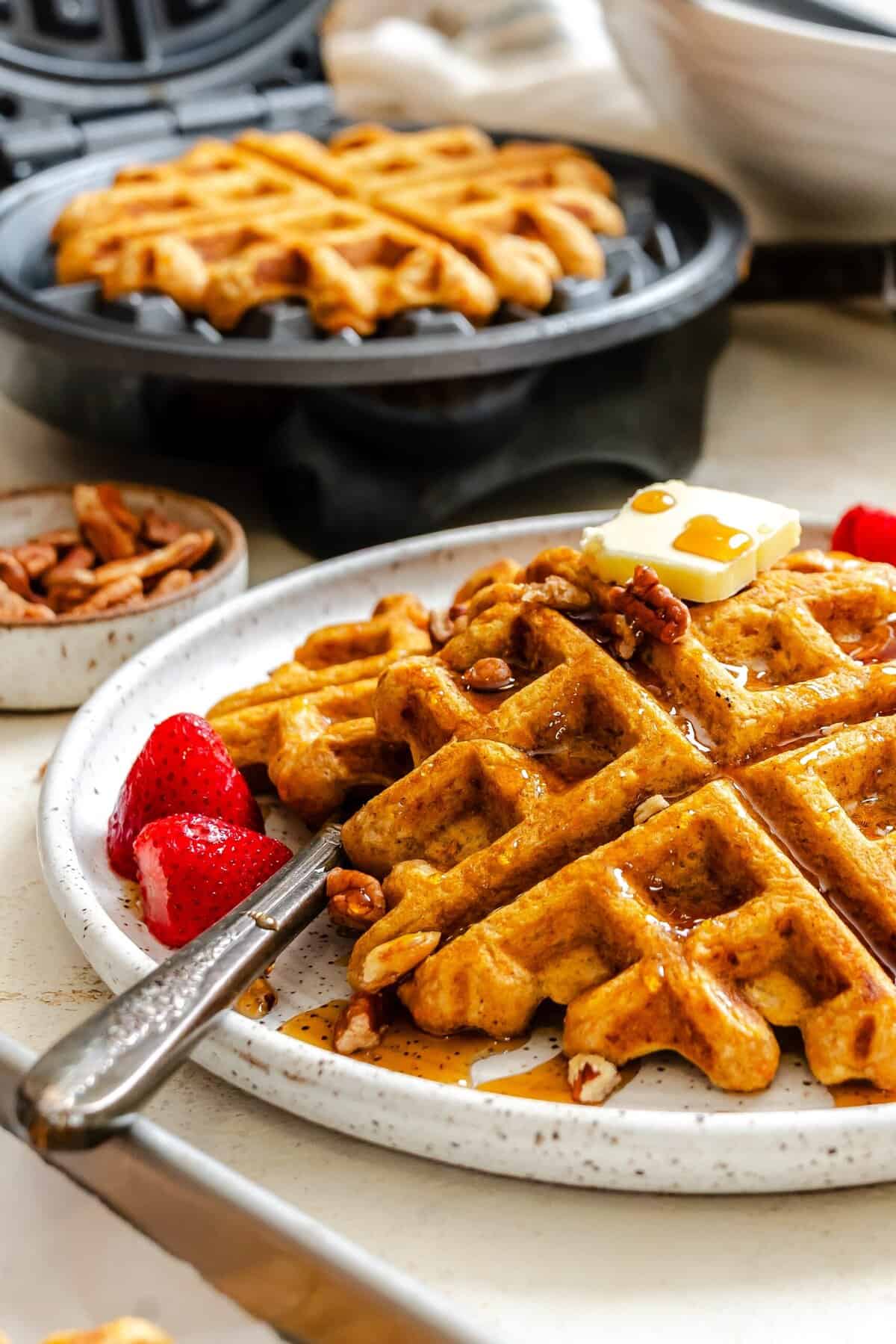 A pile of waffles with pecans and strawberries with the waffle iron in the background.